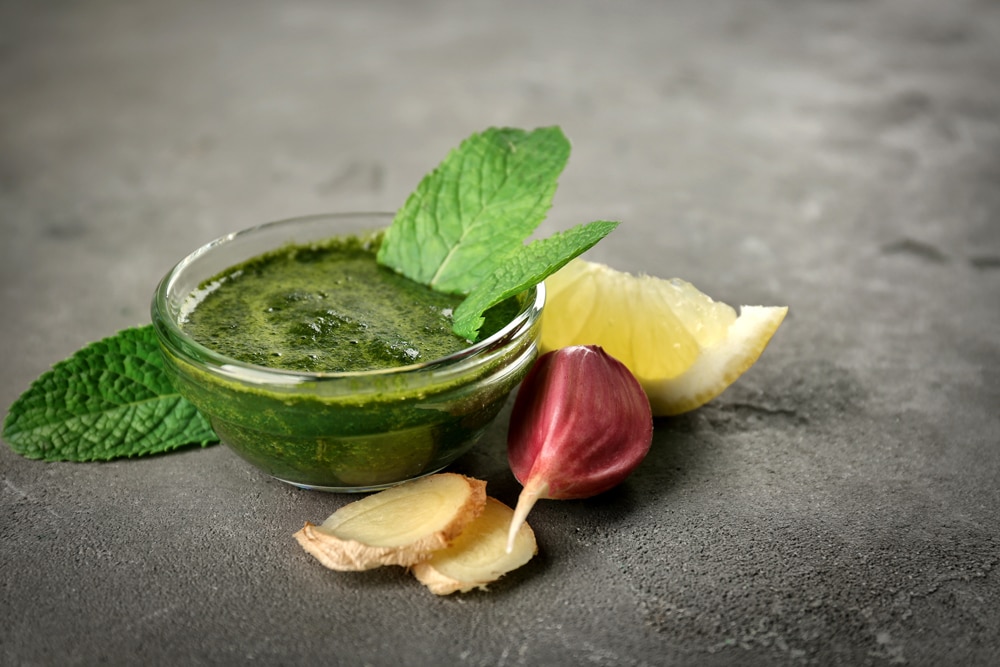 Mint chutney in a glass bowl with garlic clove, ginger slice and lemon wedge